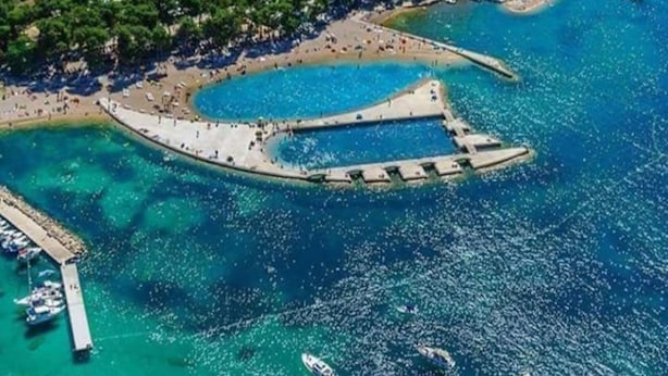 Vista aérea de una zona costera con aguas azules cristalinas, una playa de arena y un muelle con barcos.