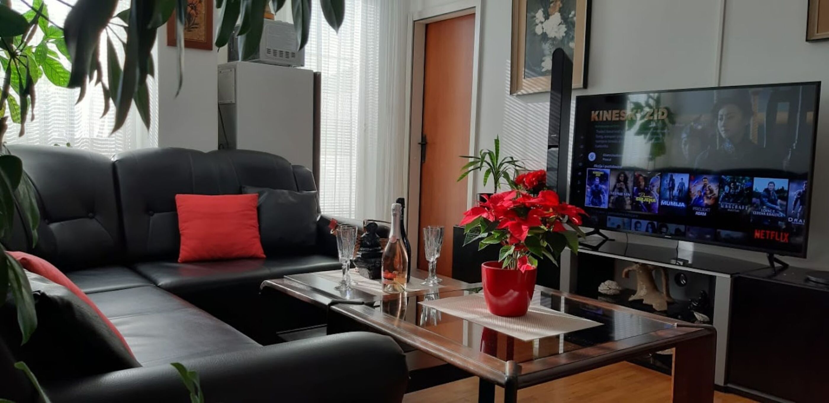 A living room with a black sofa, a red pillow, a coffee table with a vase of red flowers, and a TV displaying movie options.