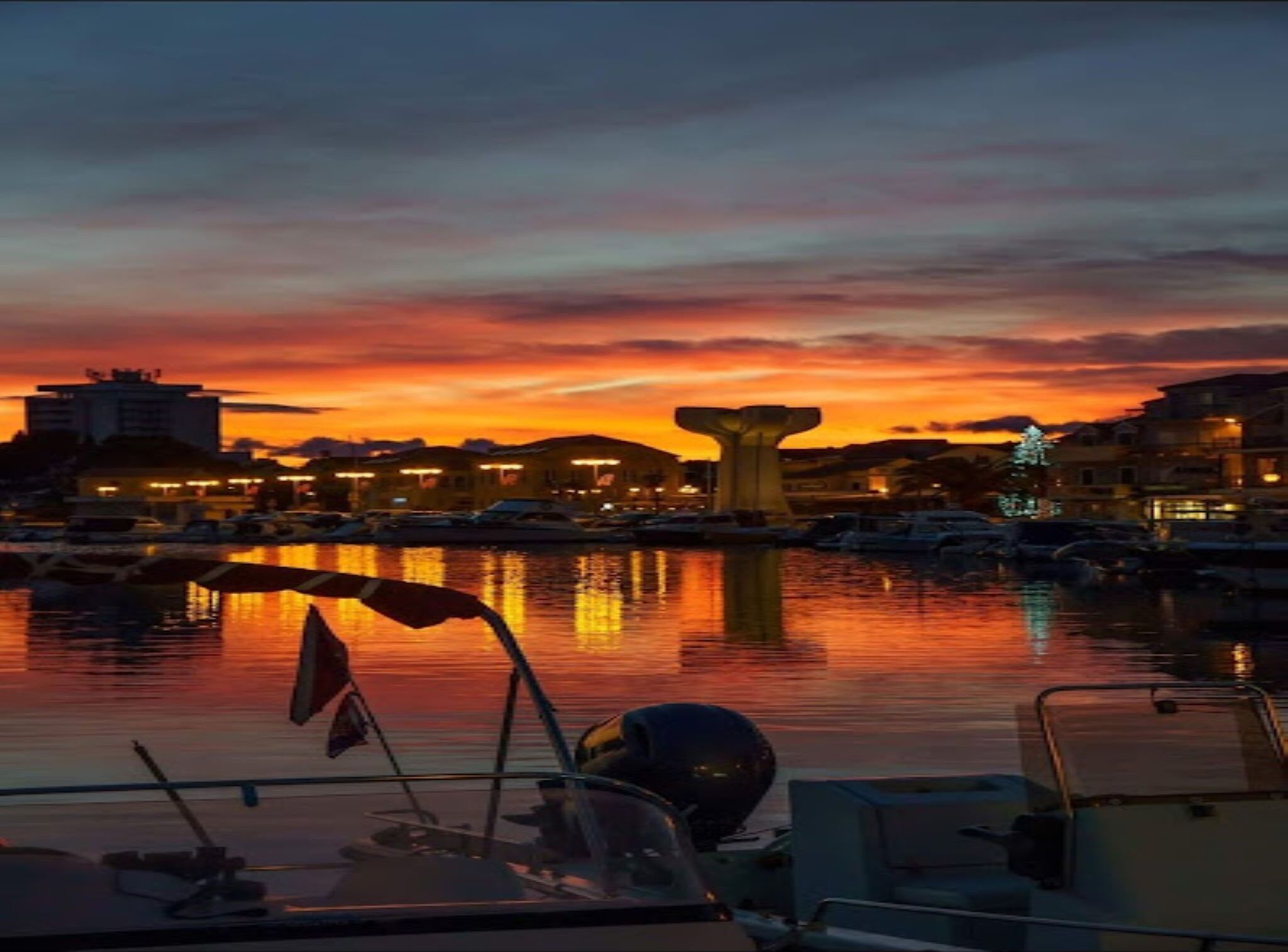 A marina at sunset with boats docked, a prominent sculpture, and buildings in the background.
