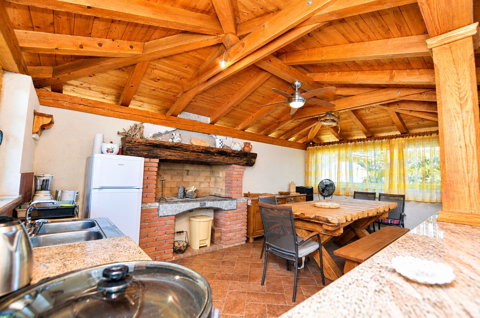 A rustic kitchen with a brick oven, wooden ceiling beams, a dining area with a table and chairs, and a refrigerator.