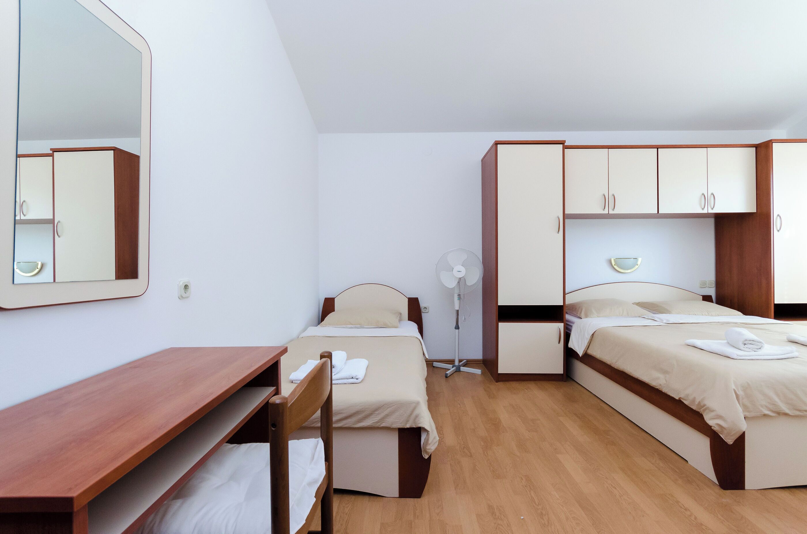 A room with two beds, a desk, a mirror, and a fan.