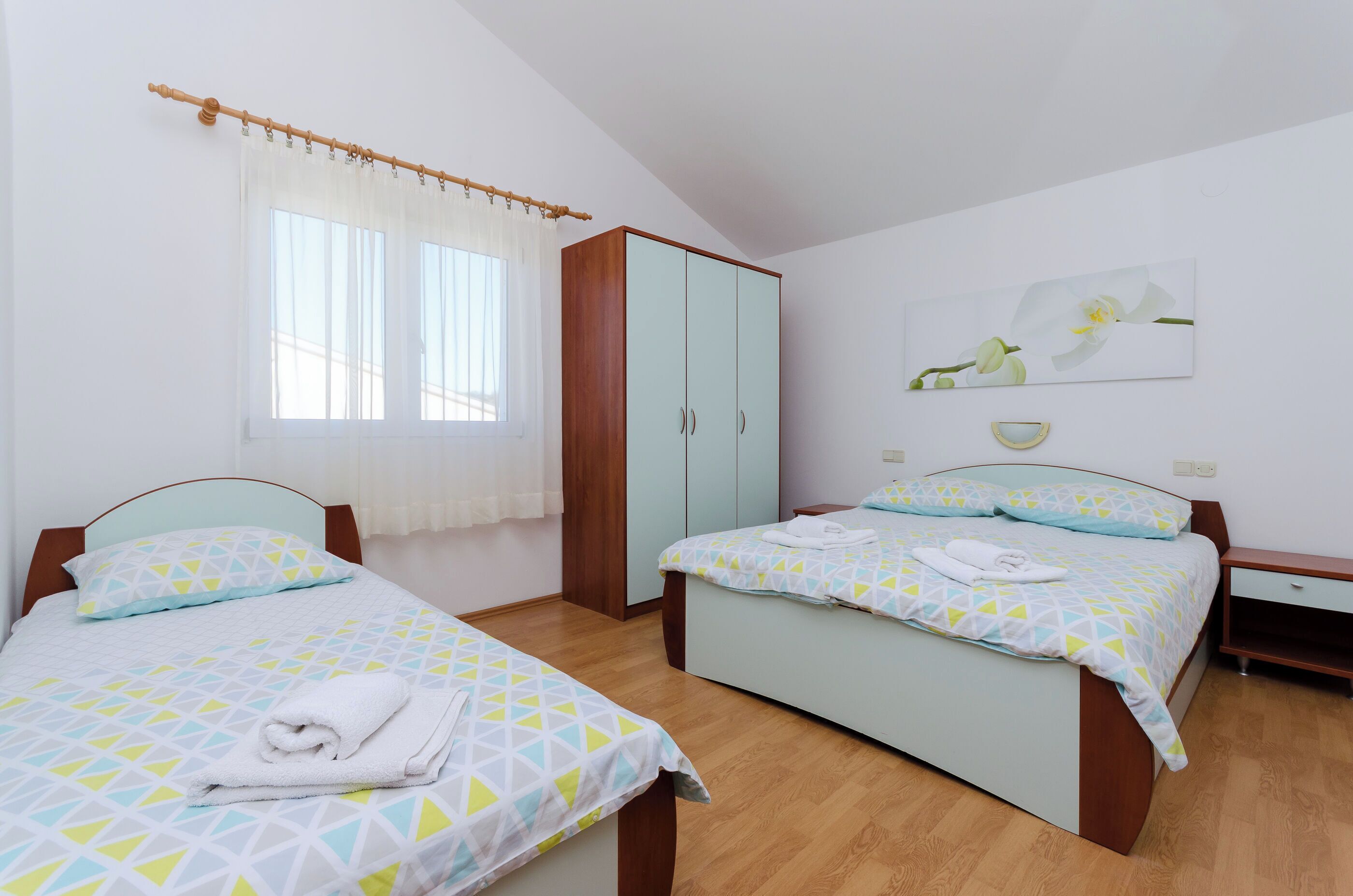 Une chambre avec deux lits, une armoire, une fenêtre avec des rideaux et un tableau représentant une orchidée accroché au mur.