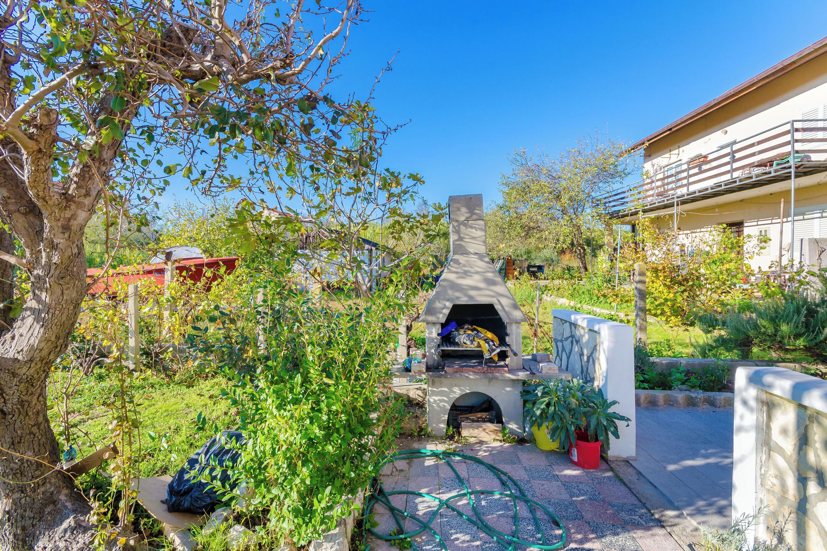 A stone-built outdoor fireplace surrounded by greenery, with a building in the background.