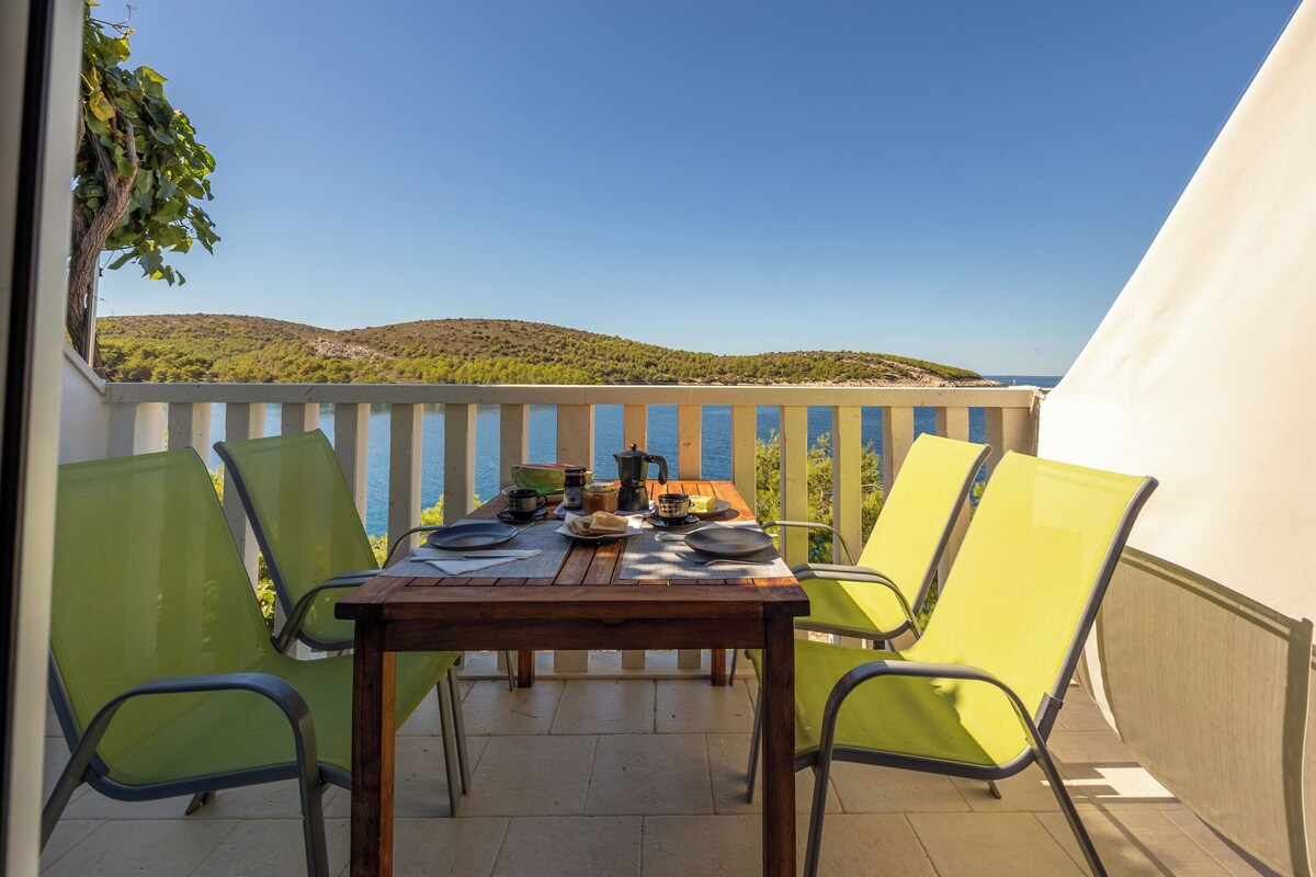 Ein Balkon mit einem Holztisch, gedeckt für zwei Personen, umgeben von grünen Stühlen und Blick auf eine Küstenlandschaft.