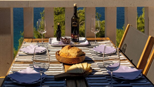 Mesa de comedor servida con copas de vino, una botella, pan y un bol, con vistas a un paisaje costero.