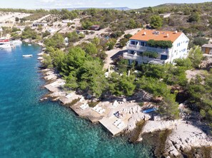 Luftaufnahme eines Küstengrundstücks mit einem großen Haus, klarem, blauem Wasser, felsiger Küstenlinie und einigen Booten.