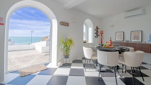 Een moderne eetruimte met een geruit vloerkleed, witte stoelen, een ronde tafel en uitzicht op de zee door een rondboogdeur.