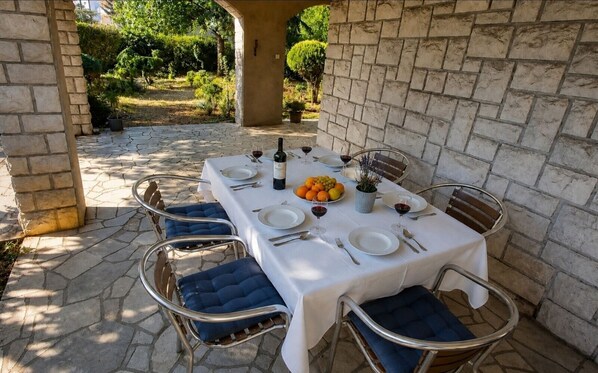 An outdoor dining area with a stone-tiled floor, a white tablecloth-covered table, blue cushioned chairs, and a fruit bowl. - Apartments Vukelic (Crikvenica)