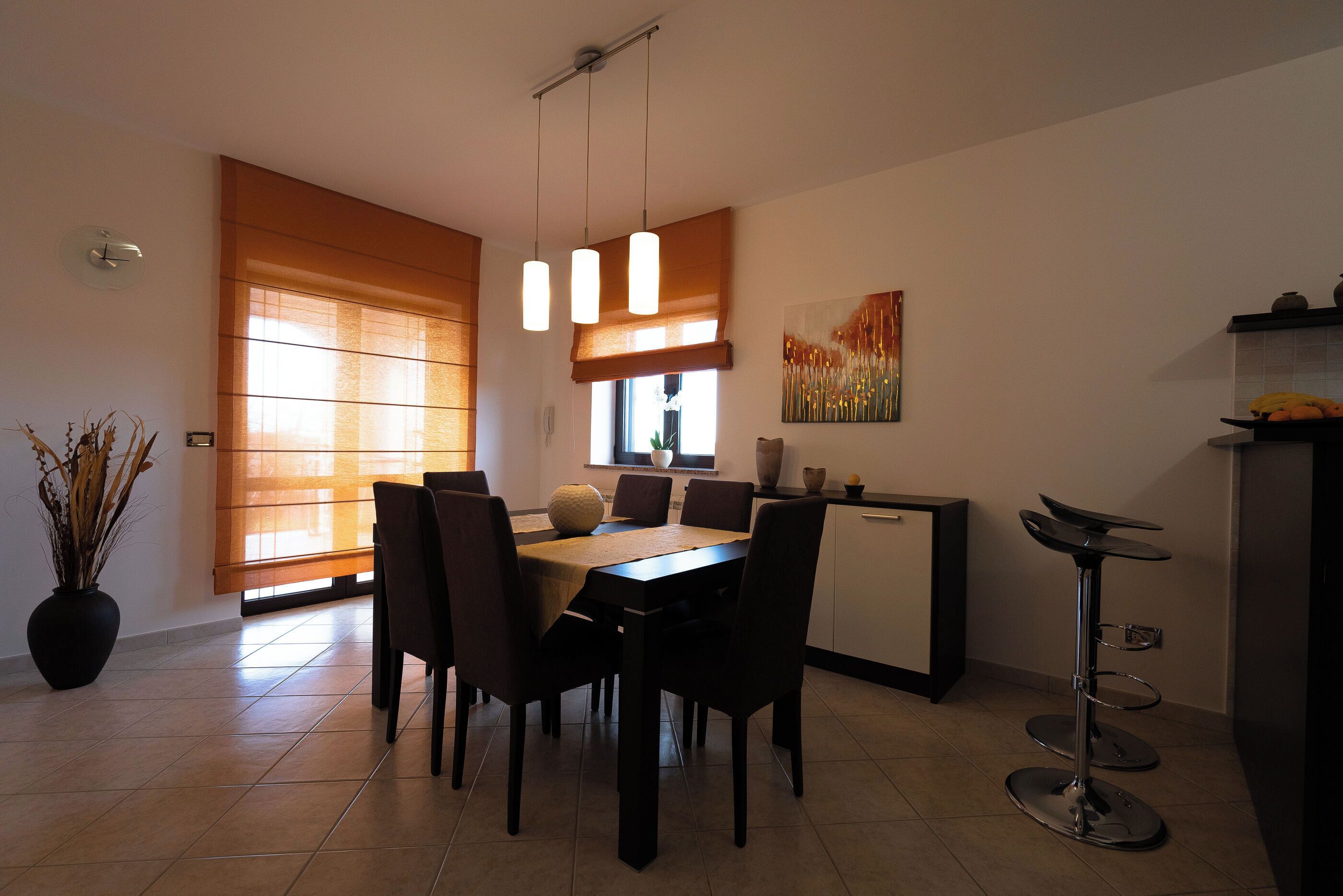 Een eetgedeelte met een tafel en stoelen, een keuken met een koelkast, en een raam met jaloezieën.