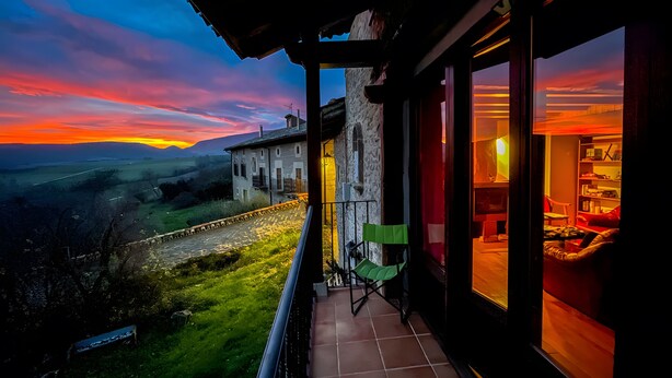 Un balcón con vista a una puesta de sol sobre un valle, un edificio de piedra y una habitación con un ventanal grande.