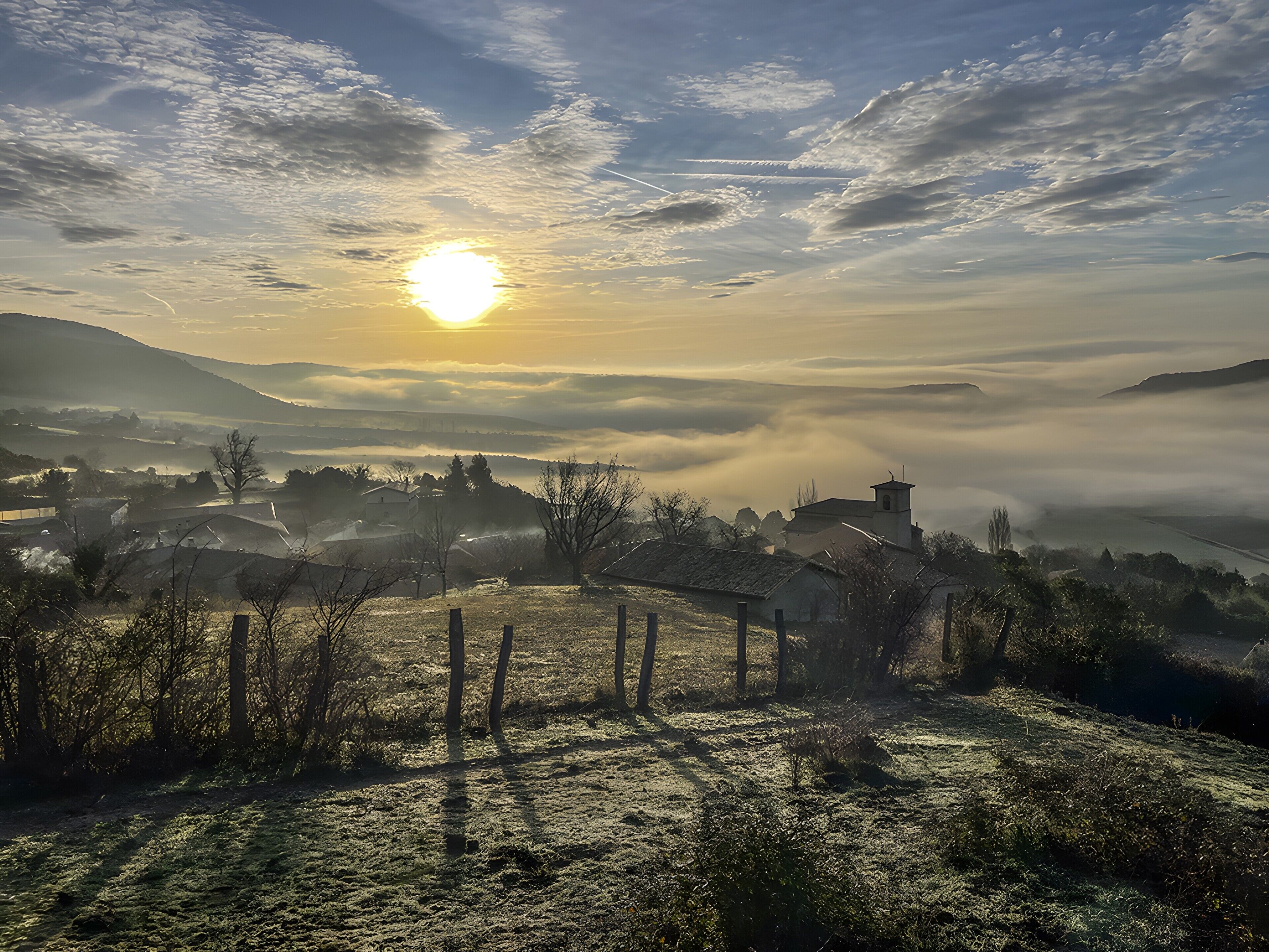 A rural landscape with a sunrise, mist-covered hills, and a small building with a tower.