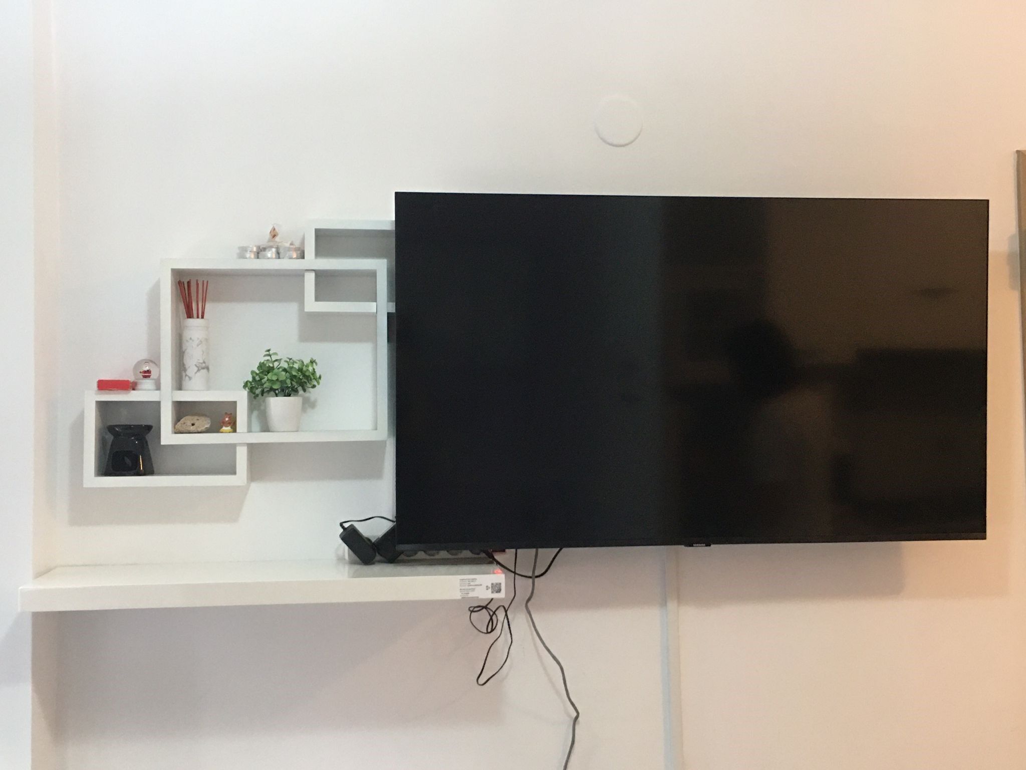 A flat-screen TV mounted on a white wall with a white shelf below it containing decorative items.