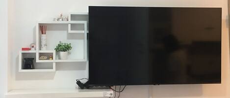 Un televisor de pantalla plana montado en una pared blanca, con un estante blanco debajo que contiene objetos decorativos.