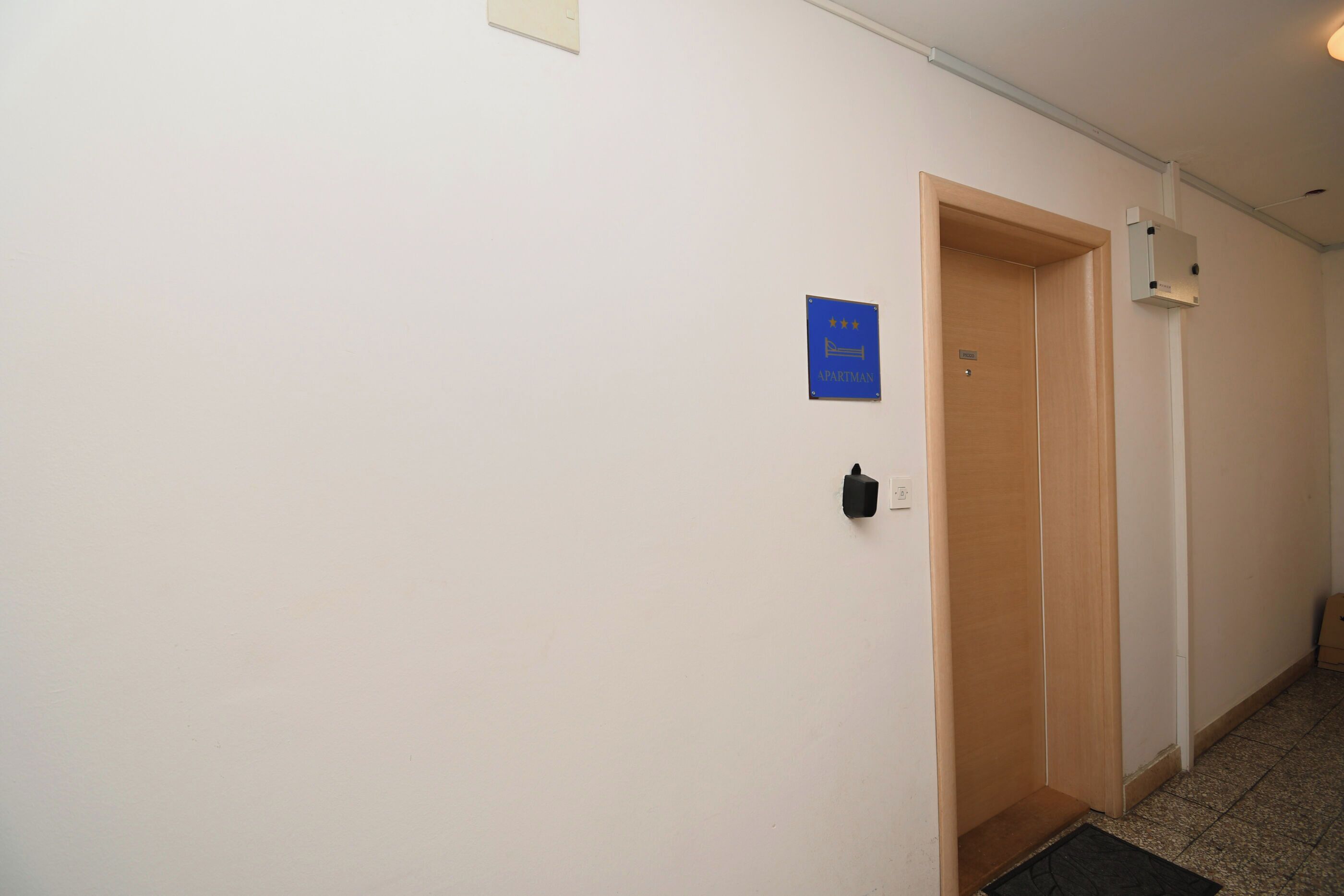 Un couloir avec une porte en bois, un panneau bleu et un interrupteur sur le mur.