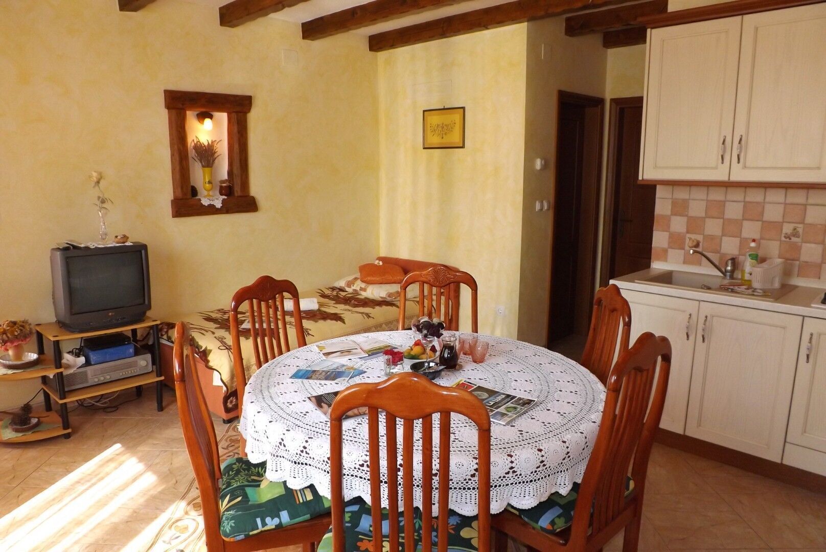 A dining area with a round table covered by a lace tablecloth, surrounded by wooden chairs. A television is placed on a wooden stand to the left.