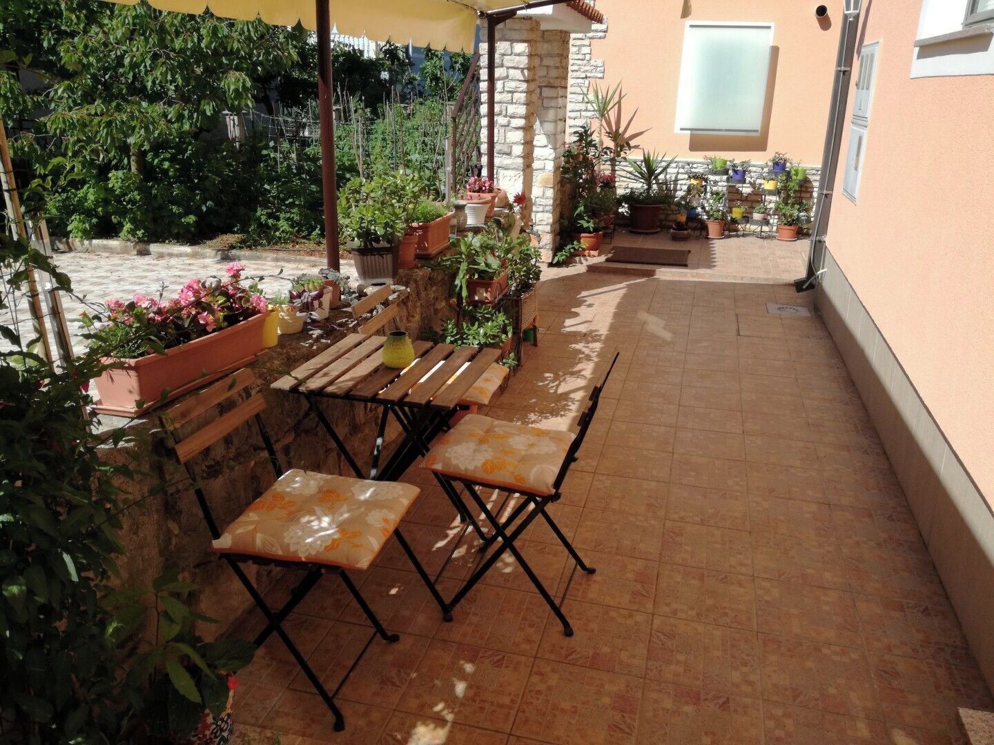 An outdoor patio with wooden tables and chairs, surrounded by potted plants and a stone wall.