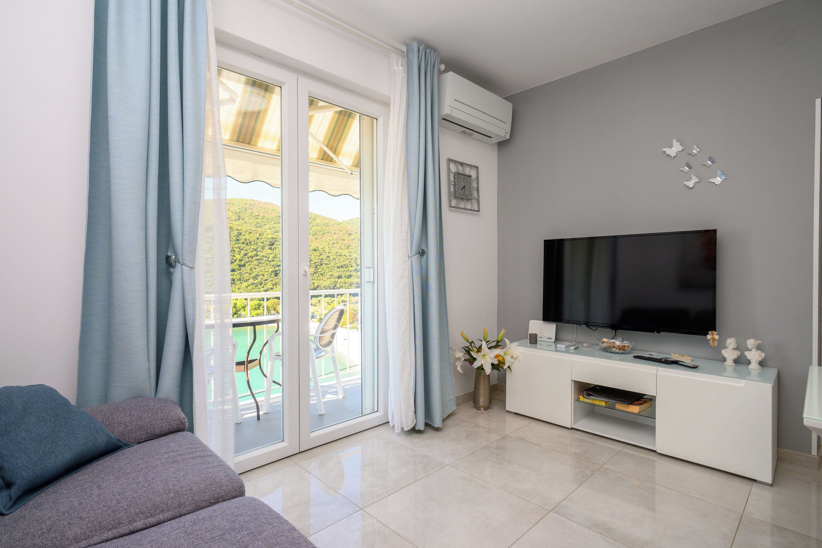 A modern living room with a flat-screen TV, a sofa, a dining area with chairs, and a balcony with a view of greenery.