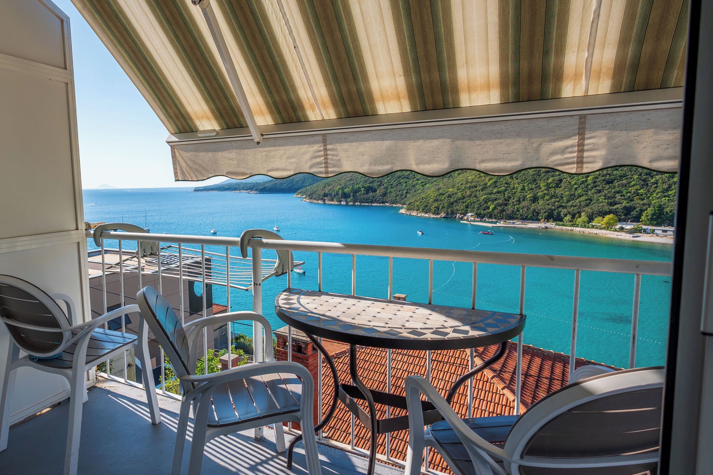 A balcony with a view of a turquoise sea, a small round table, and two chairs.