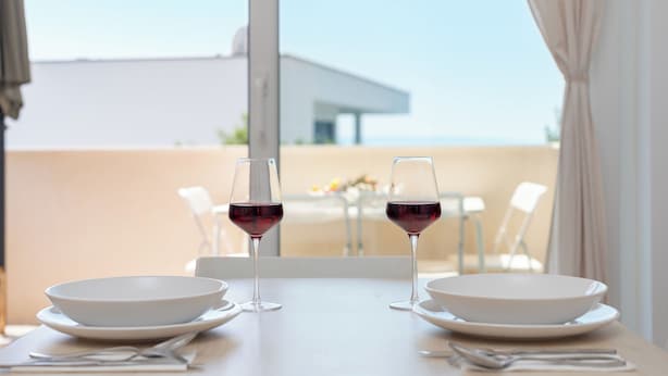 A dining table with two white bowls and two wine glasses on white plates, set against a backdrop of a clear sky and a building. - Apartments Marina (Novalja)