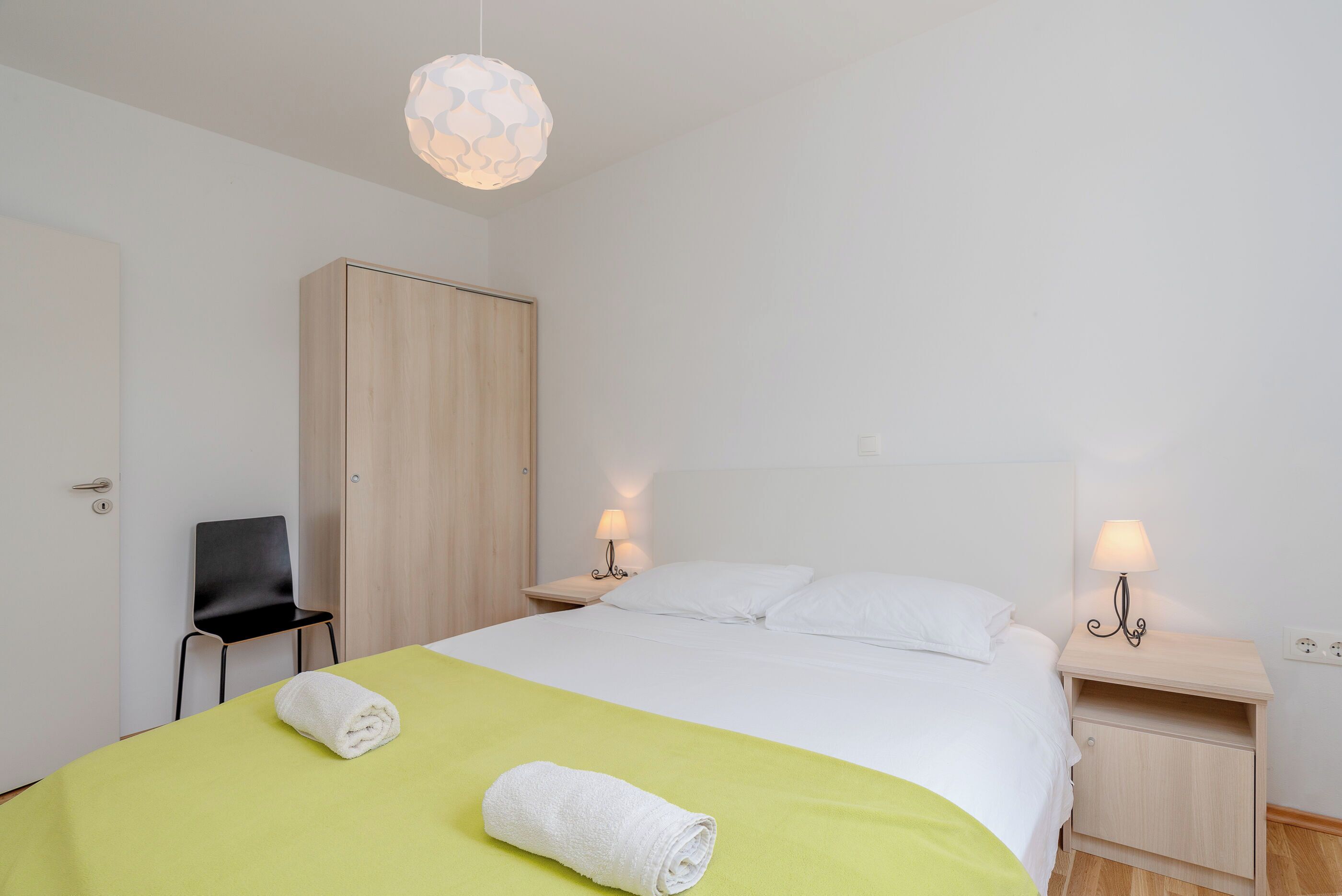 A hotel room with a bed, a chair, a wardrobe, and a nightstand with a lamp.