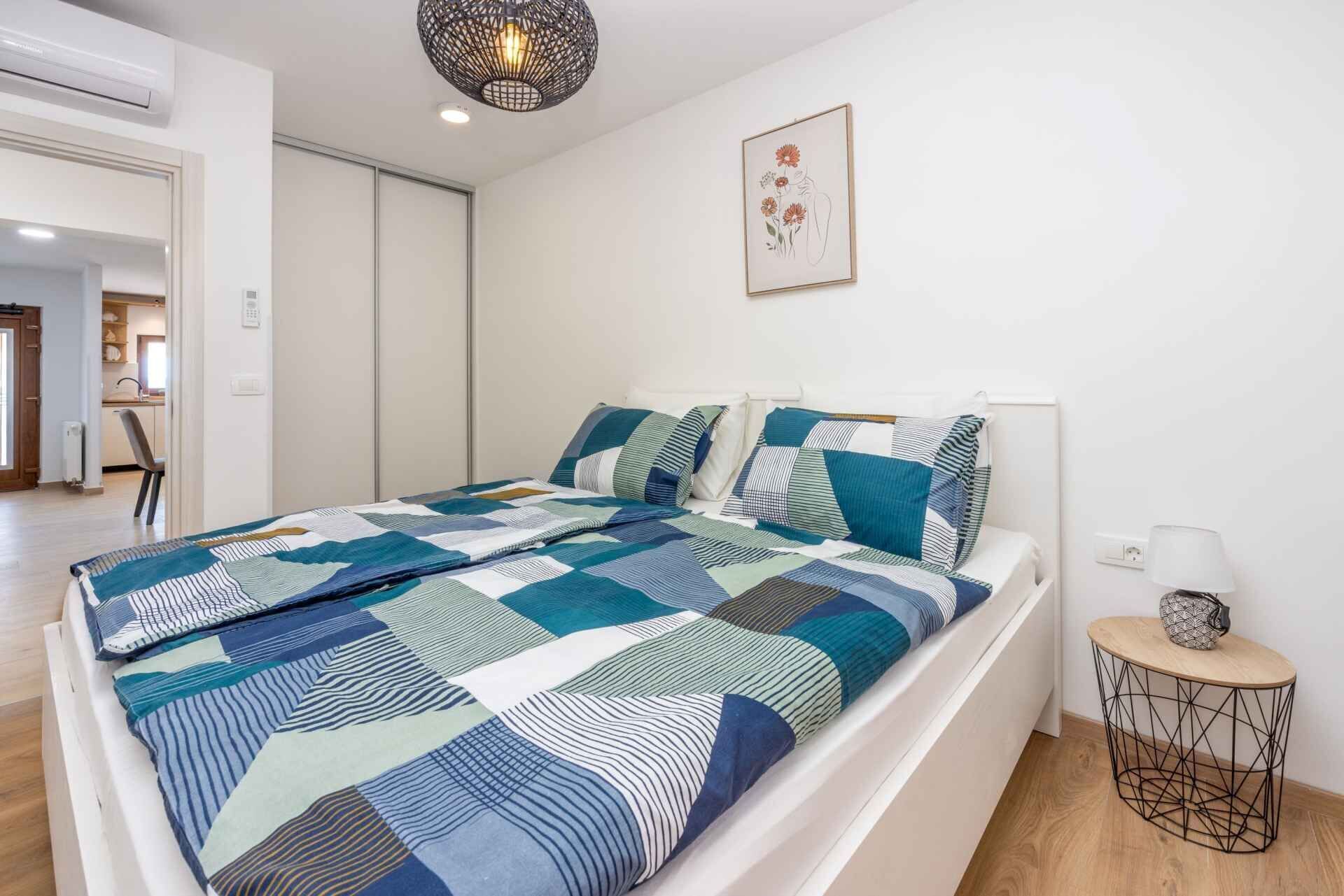 Quarto moderno com uma cama grande, uma mesinha de cabeceira, um lustre e uma obra de arte emoldurada na parede.