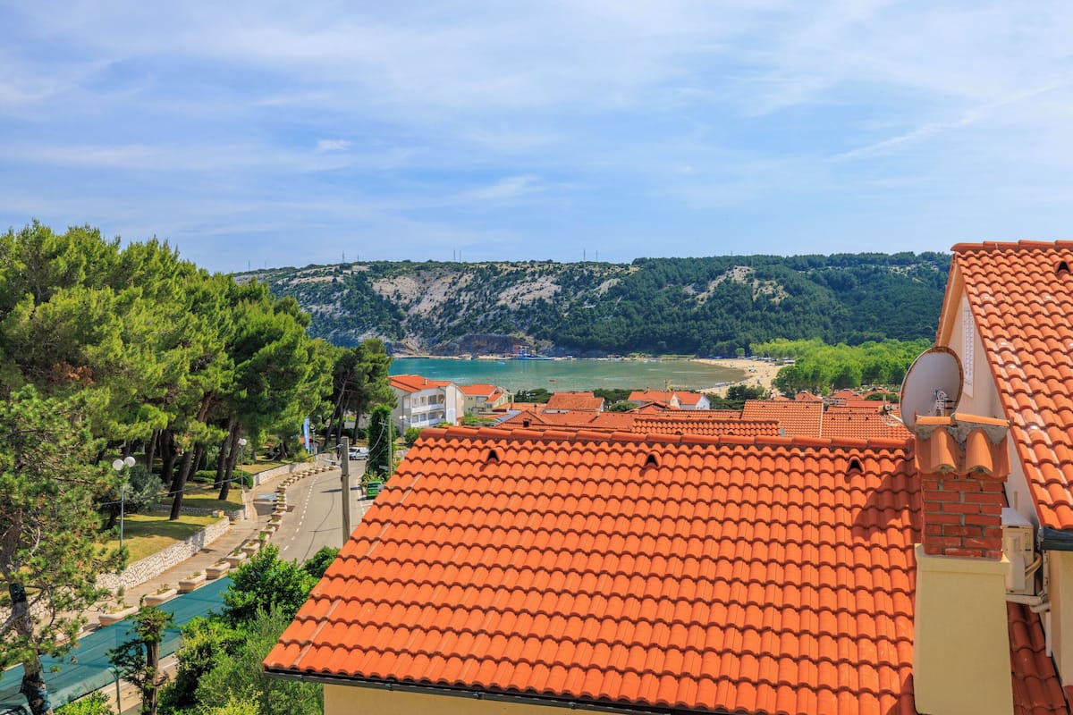 A coastal view with terracotta-tiled roofs, a swimming pool, and a beach in the distance.
