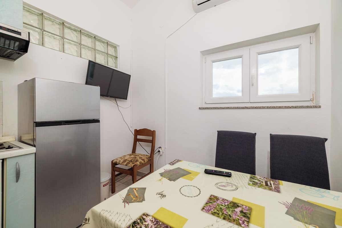 A compact kitchen with a refrigerator, a dining table with a floral tablecloth, two chairs, a window with blinds, and a wall-mounted television.