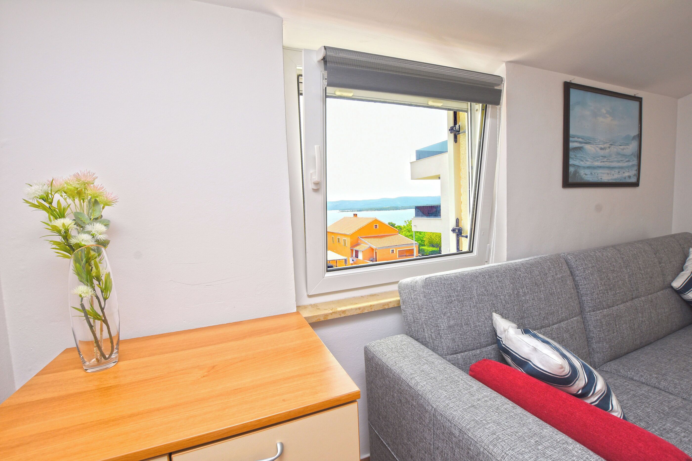 A room with a grey sofa, a wooden side table, a vase with flowers, a window with a view of buildings and water, and a framed picture on the wall.