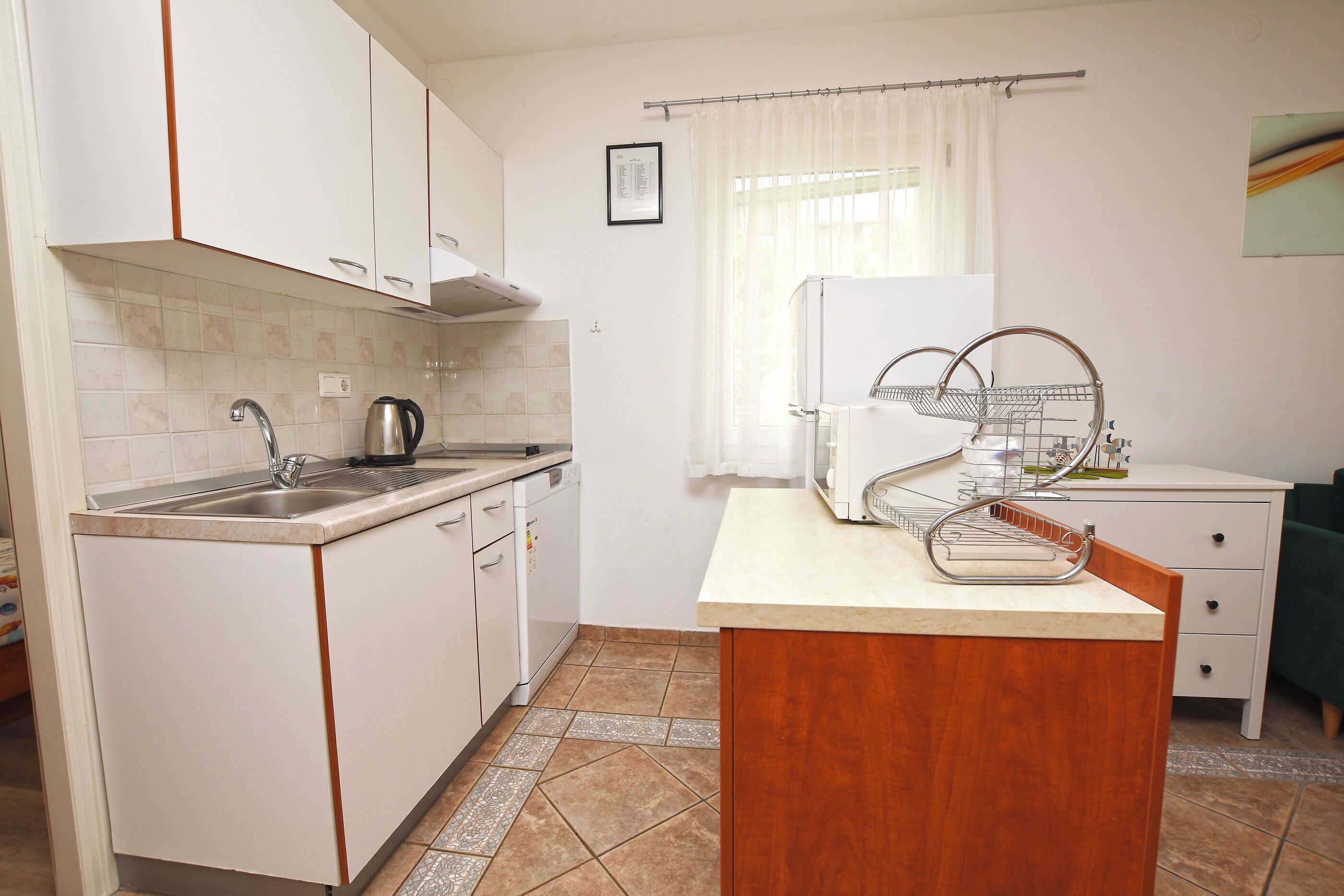 Cocina compacta con armarios blancos, un fregadero, un hervidor y una pequeña isla con microondas.