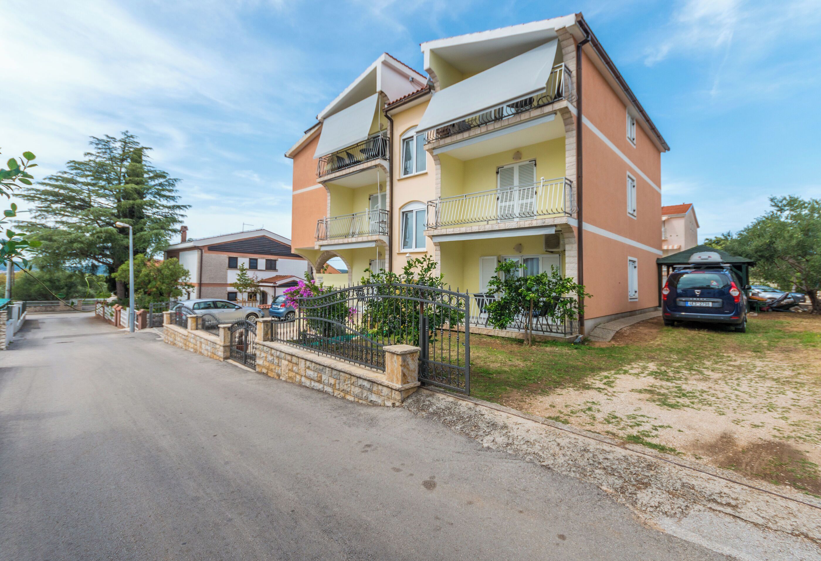 A multi-story residential building with balconies, a gated driveway, and parked cars.