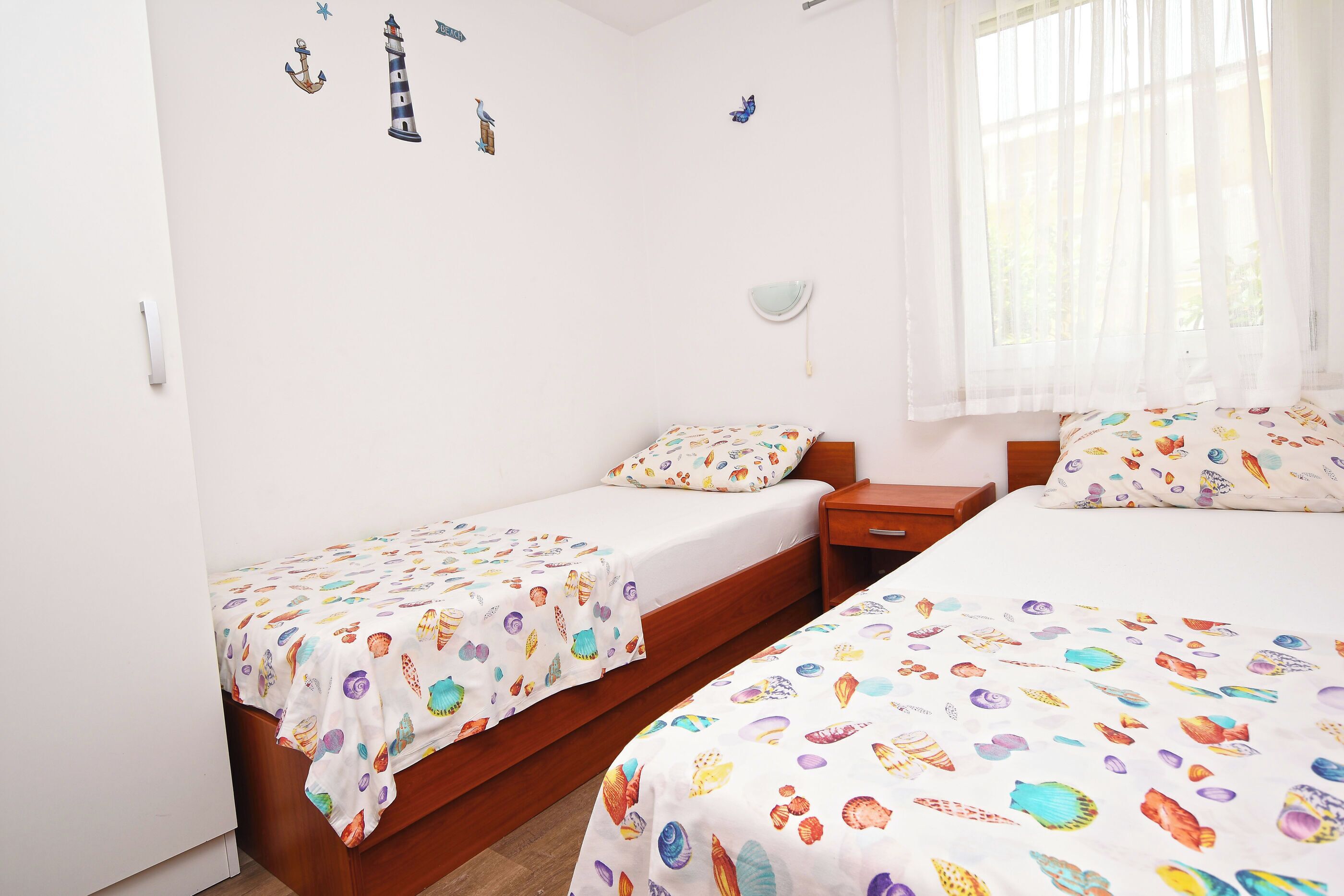 A bedroom with two single beds, a bedside table, a nightstand, and a window with sheer curtains.
