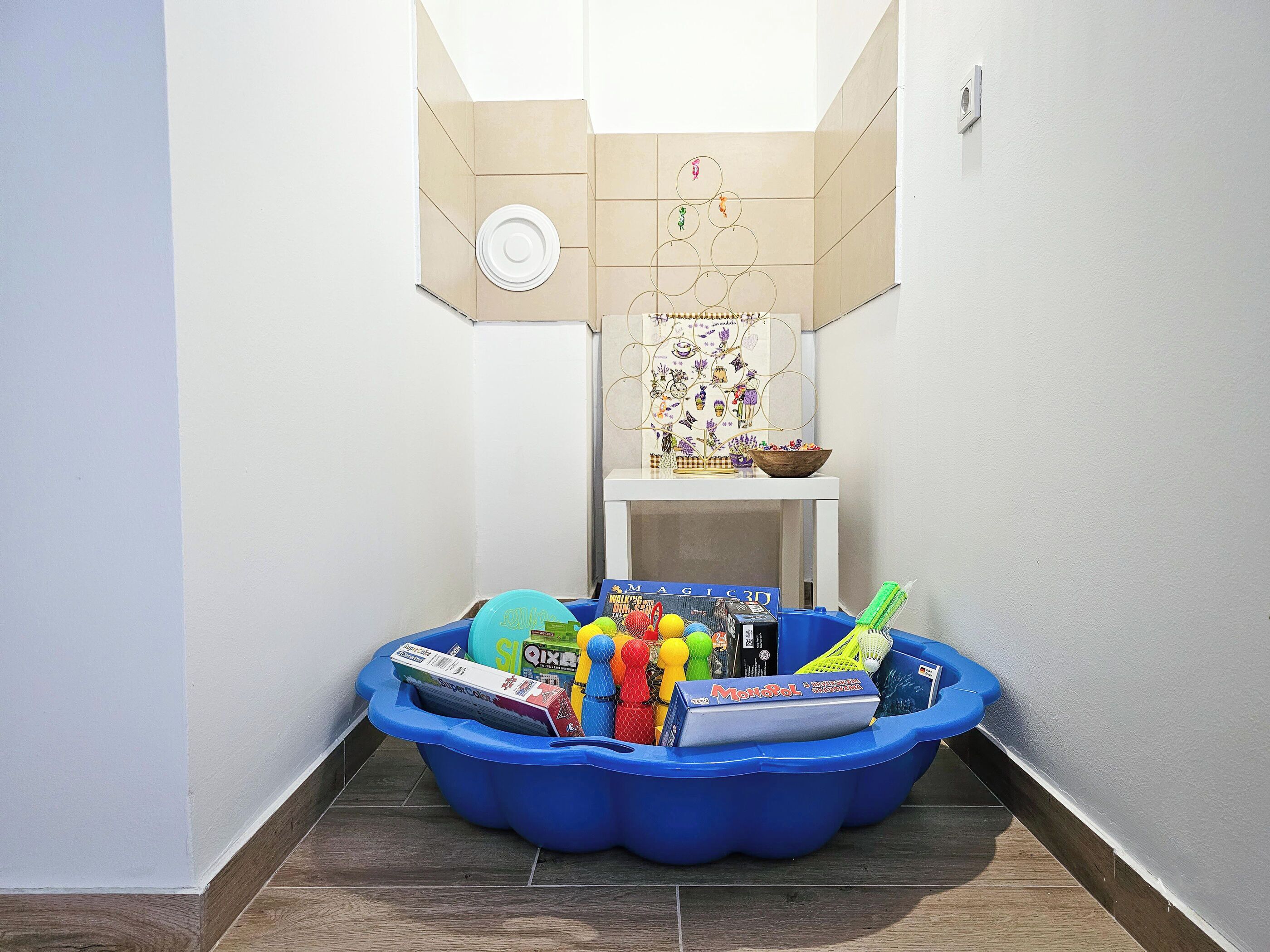 A corner of a room with a blue plastic storage bin filled with toys and games, a white shelf with a decorative item, and a wall with a circular cutout.
