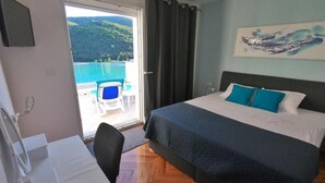 Un dormitorio con cama, escritorio y silla. Hay una ventana con cortinas y vistas a un cuerpo de agua y colinas.