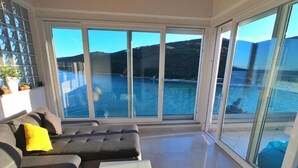 Una sala de estar moderna con un sofá gris, una mesa de centro blanca y grandes ventanas que ofrecen vistas a un cuerpo de agua y colinas.