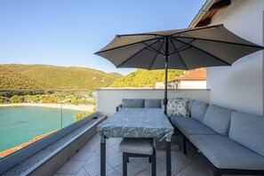 A balcony with a table and a sofa, overlooking a coastal area with a view of the sea and hills. - Apartment Beautifull View (Labin)