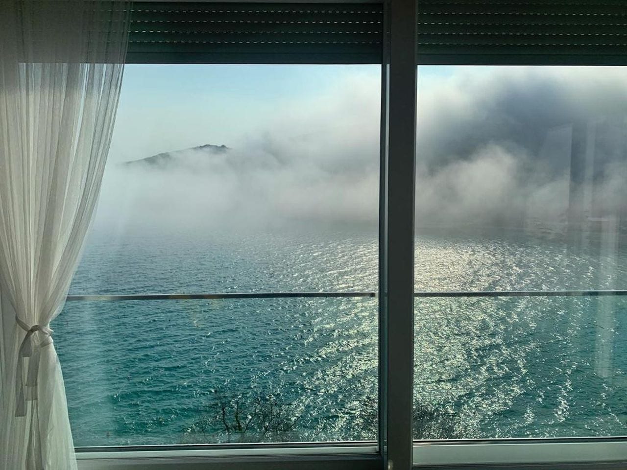 Une vue par une fenêtre ornée de rideaux légers, donnant sur une étendue d’eau avec, au loin, une masse terrestre partiellement dissimulée par les nuages.