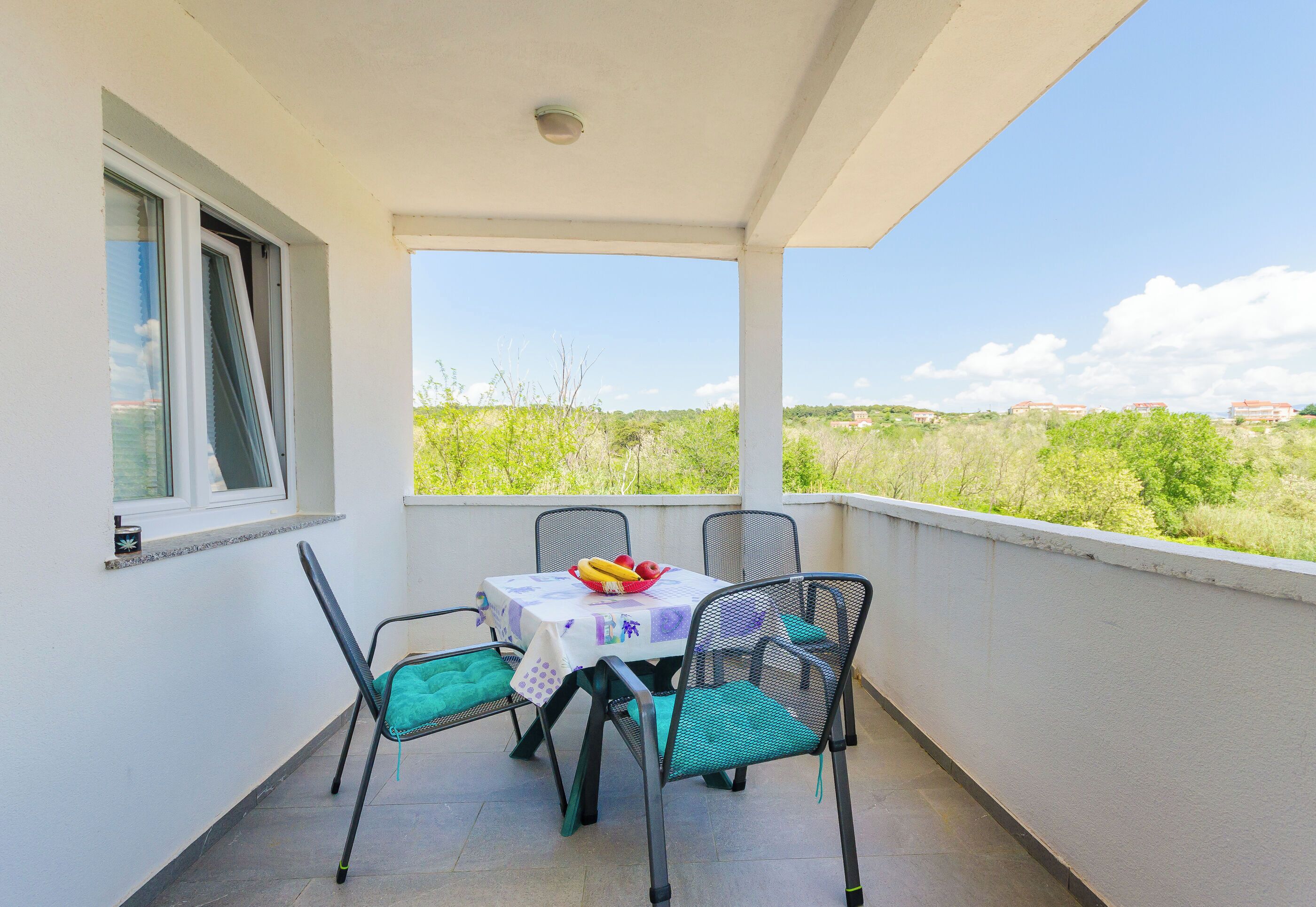 Un balcón con mesa y sillas, una ventana y vistas a zonas verdes y edificios.