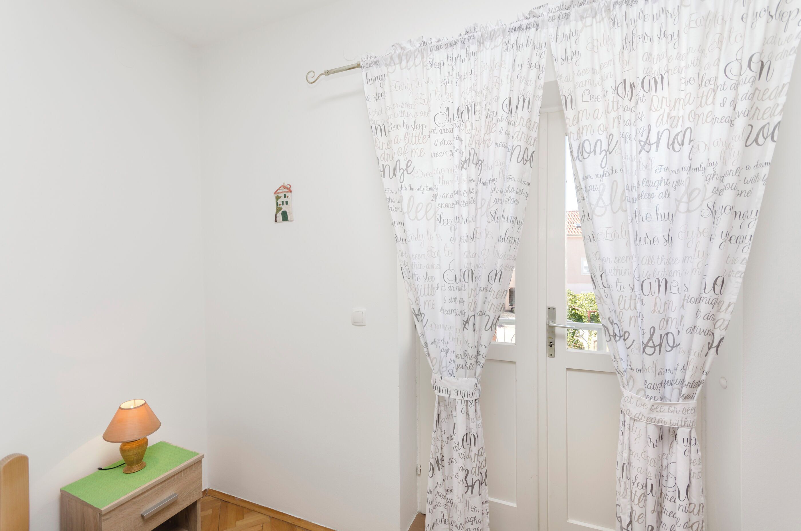 A room with a wooden nightstand, a lamp, and a white door with a curtain featuring motivational quotes.