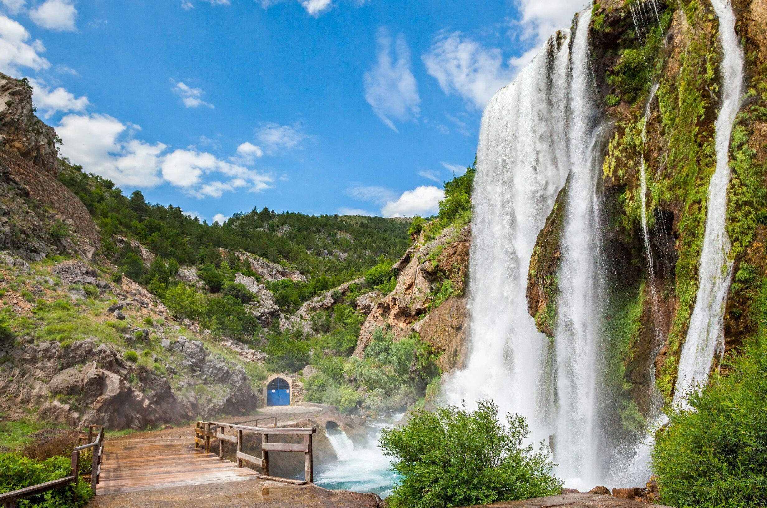Eine Holzbrücke führt zu einem Wasserfall, im Hintergrund eine blaue Tür.