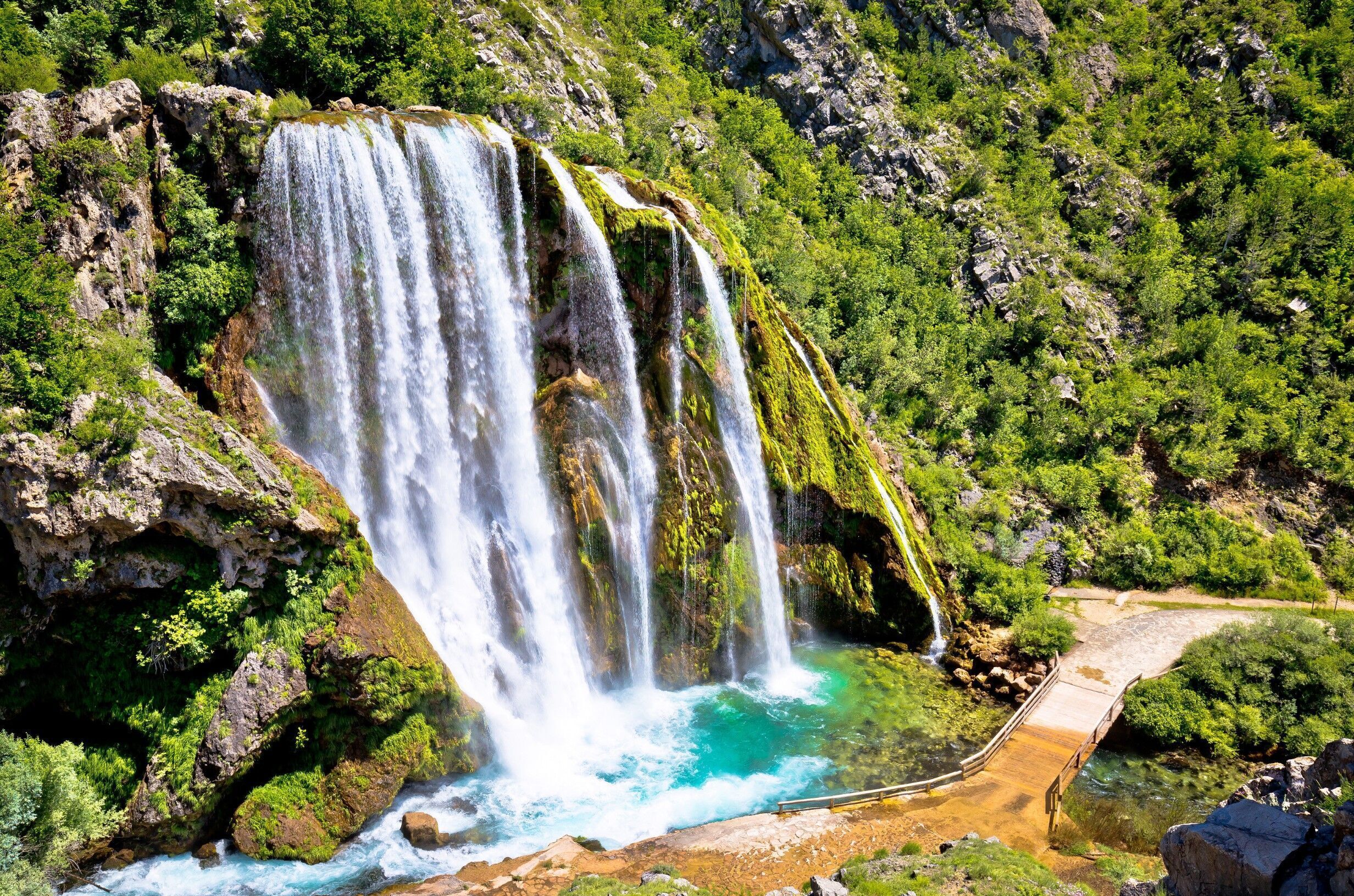 Uma cascata desce por um penhasco rochoso, cercada por vegetação exuberante.