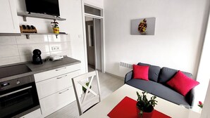 A modern kitchen with a TV, a coffee machine, and a microwave. There is a grey sofa with red cushions and a red table with a plant. - Apartment Oliva (Knin)