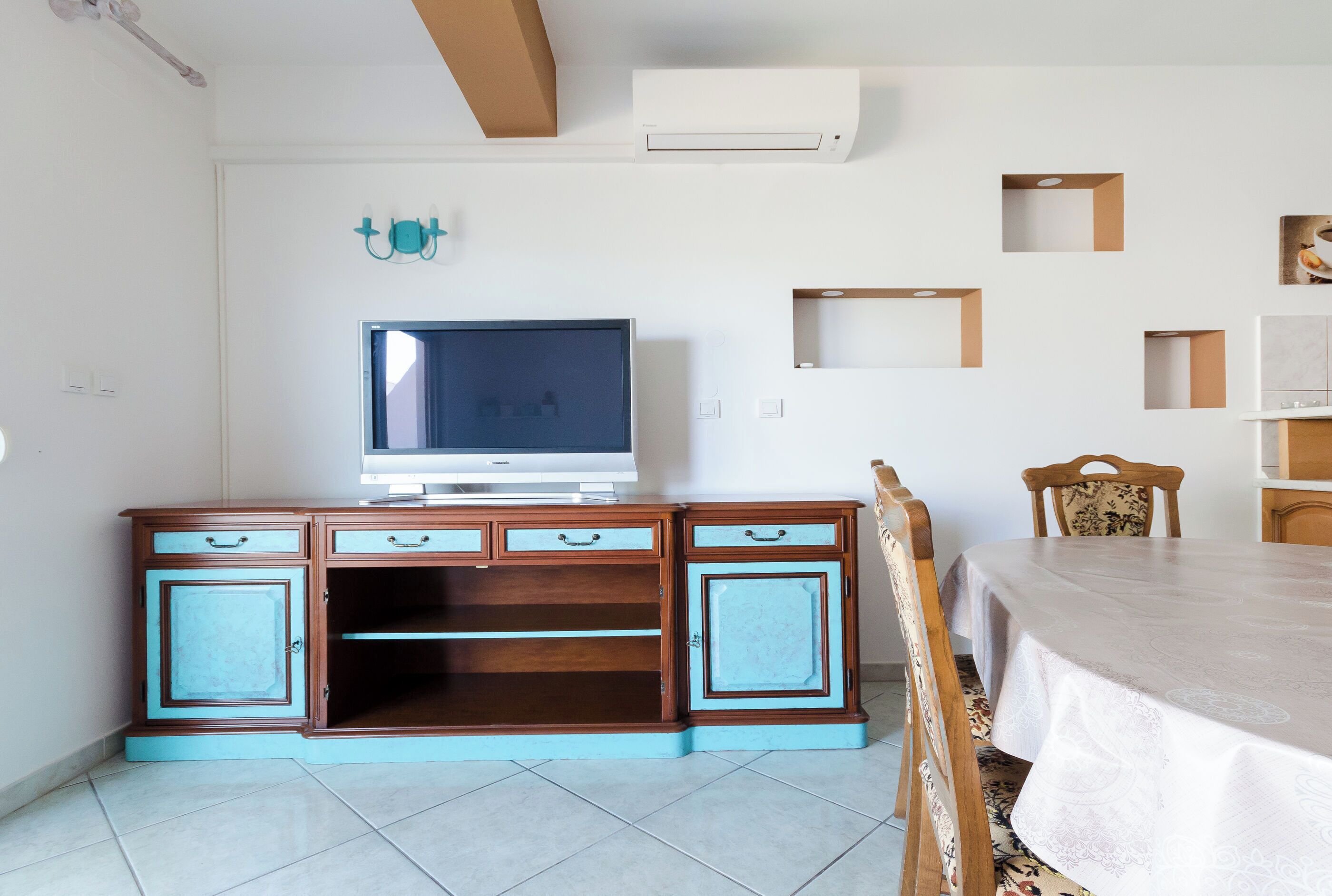 Ein Zimmer mit Fernseher auf einer Holzkommode, einem Esstisch mit Stühlen und einer Wandklimaanlage.