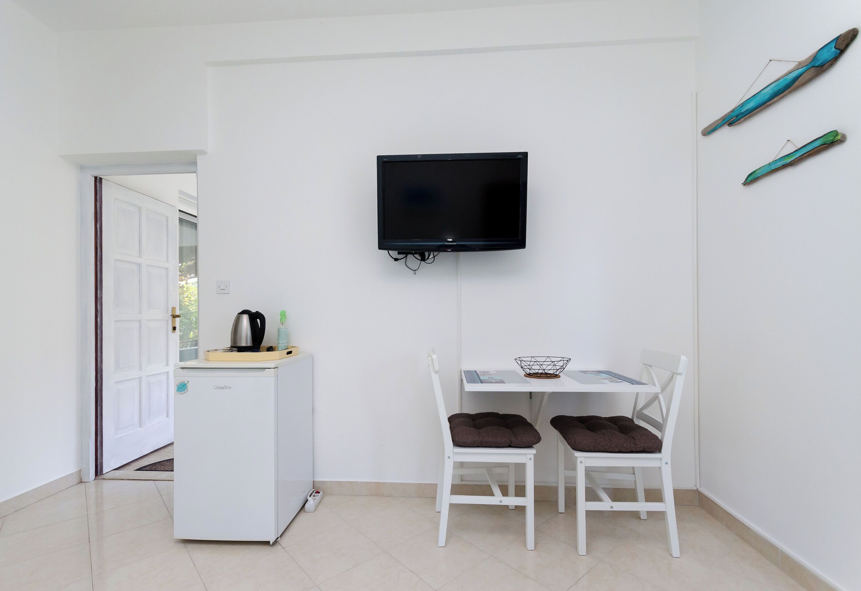 Une petite pièce propre, avec un réfrigérateur blanc, une table et deux chaises, un téléviseur à écran plat fixé au mur et une porte donnant sur une autre pièce.