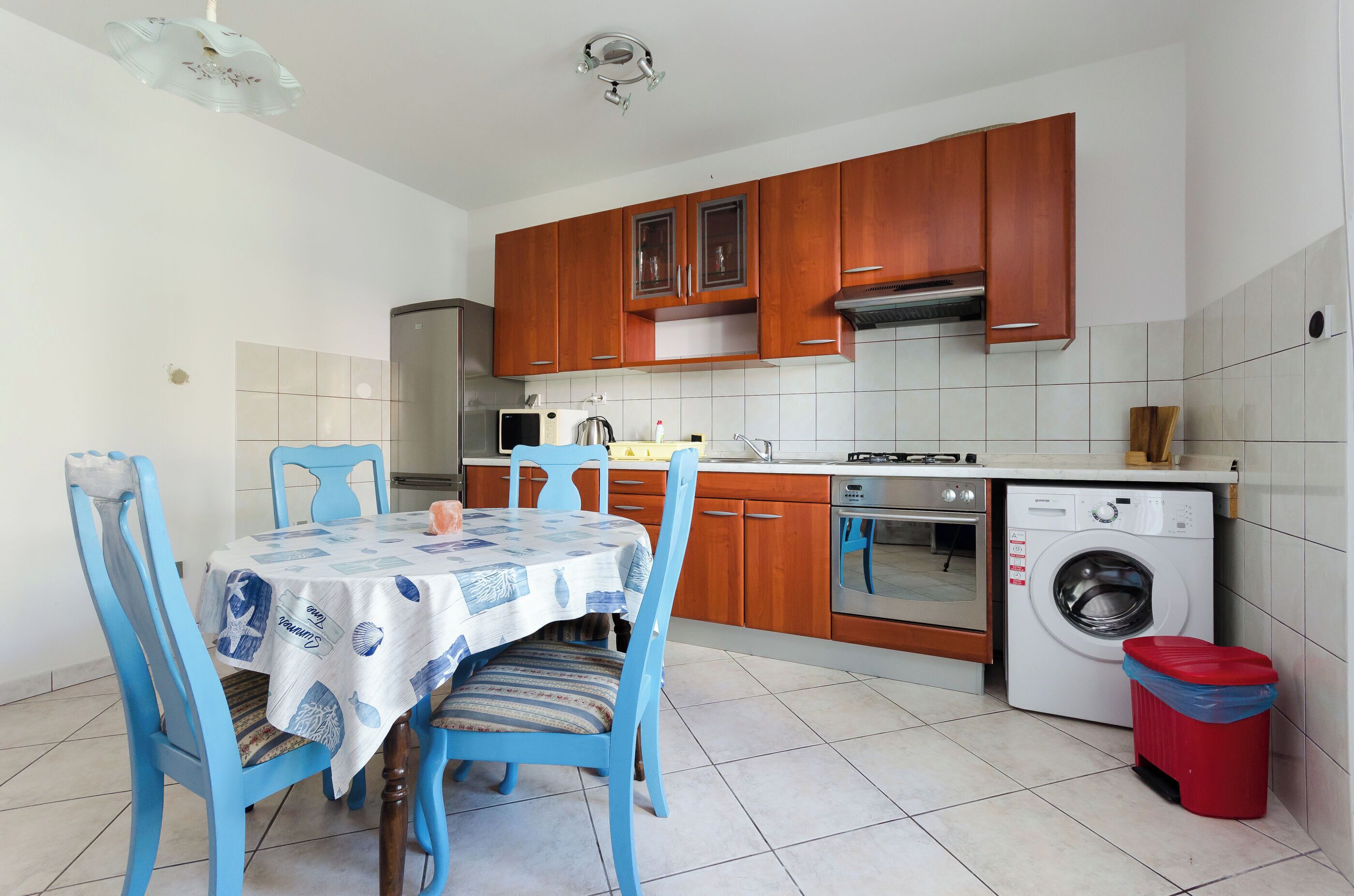 A compact kitchen with wooden cabinets, a built-in oven, a washing machine, and a dining table with chairs.