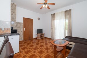 A modern living room with a wooden floor, a sofa, a glass-top coffee table, a television on a wooden stand, a ceiling fan, and a window with sheer curtains. - Apartments Ivica (Vir)