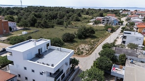 Un edificio moderno con tetto piatto, un piccolo patio con posti a sedere all'aperto e vista su una zona residenziale con case e una zona boschiva sullo sfondo.