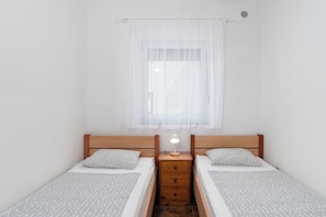 Una camera doppia con due letti singoli, un comodino con una lampada, una finestra con tende leggere e una bocchetta di ventilazione a muro.