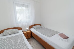 A bedroom with two single beds, a bedside table with a lamp, and a window with sheer curtains. - Apartments Ivica (Vir)
