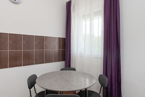 Ein Zimmer mit einem runden Tisch, vier Stühlen, einer Wanduhr und einem Fenster mit violetten Vorhängen.