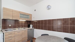 A compact kitchen with wooden cabinets, a built-in dishwasher, a coffee maker, and a round table with chairs. - Apartments Ivica (Vir)